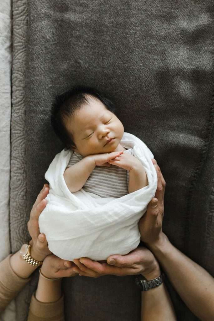 Newborn baby swaddled in a white blanket, lying on a gray surface, gently cradled by two pairs of adult hands.