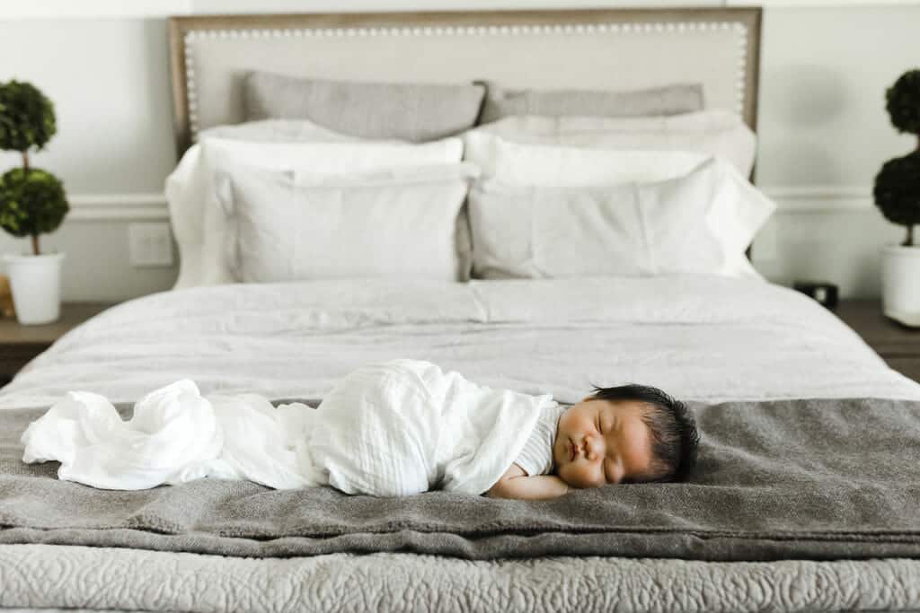 A baby wrapped in a white blanket sleeps on a gray blanket atop a neatly made bed with white pillows and potted plants in the background.