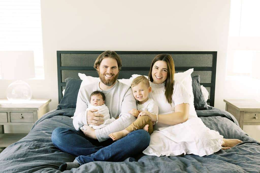 A family of four sits on a bed; the father holds a baby, while the mother and young son sit beside them, all smiling at the camera.