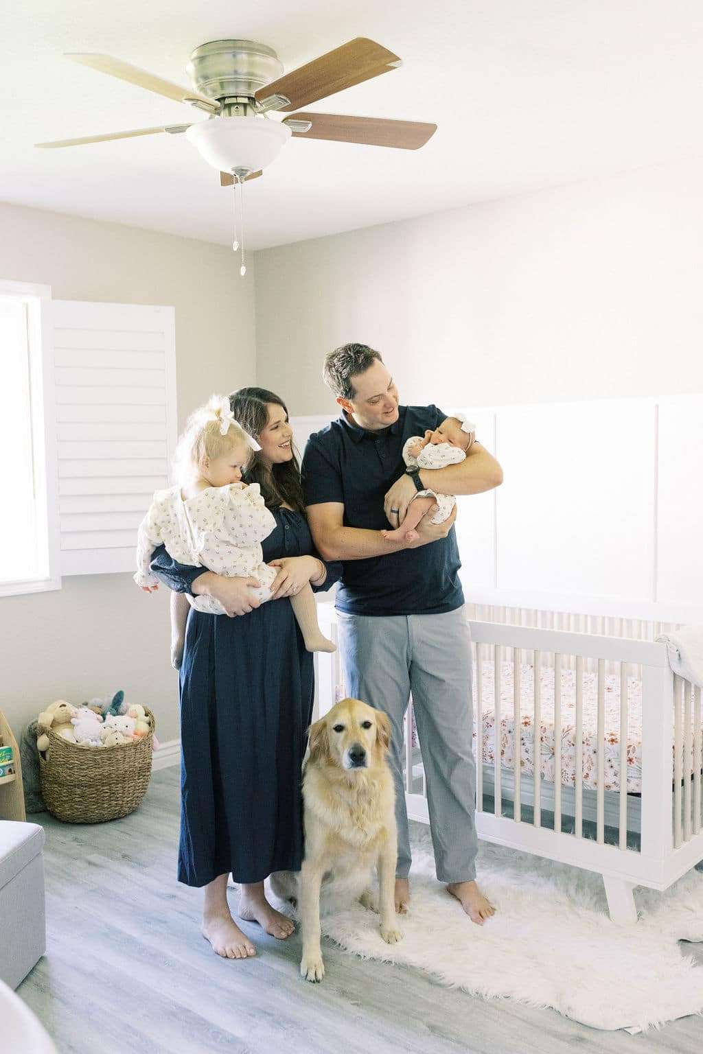 A family of four with a dog stands in a bright nursery; the father holds a baby, the mother holds a toddler, and a crib and toys are in the background.