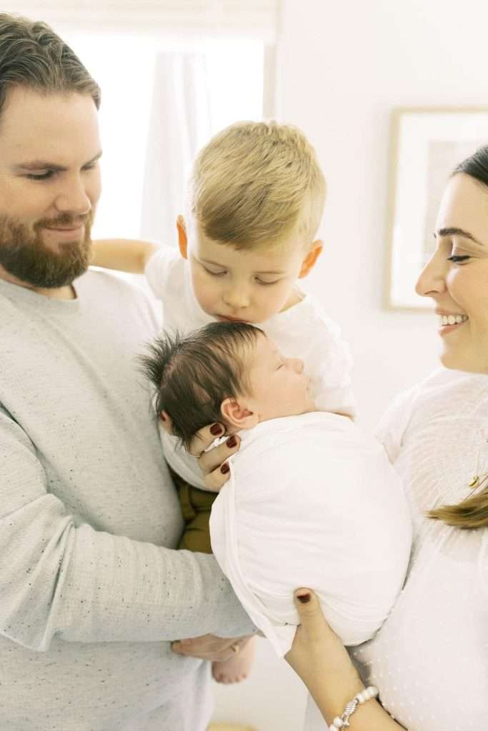Two adults and a young child admire a swaddled newborn. The child gently leans in to kiss the baby while the adults smile warmly.