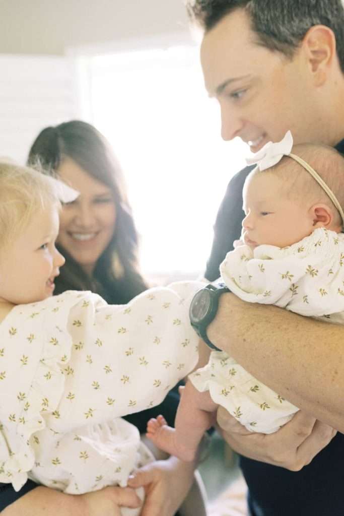 A man holds a baby while a woman holds a toddler; both children wear matching light outfits with floral patterns.