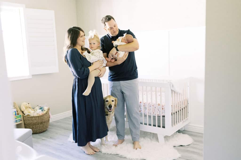 A man and woman stand in a nursery holding a baby and a toddler. A dog sits on the floor between them. There is a crib and a basket of toys in the background.