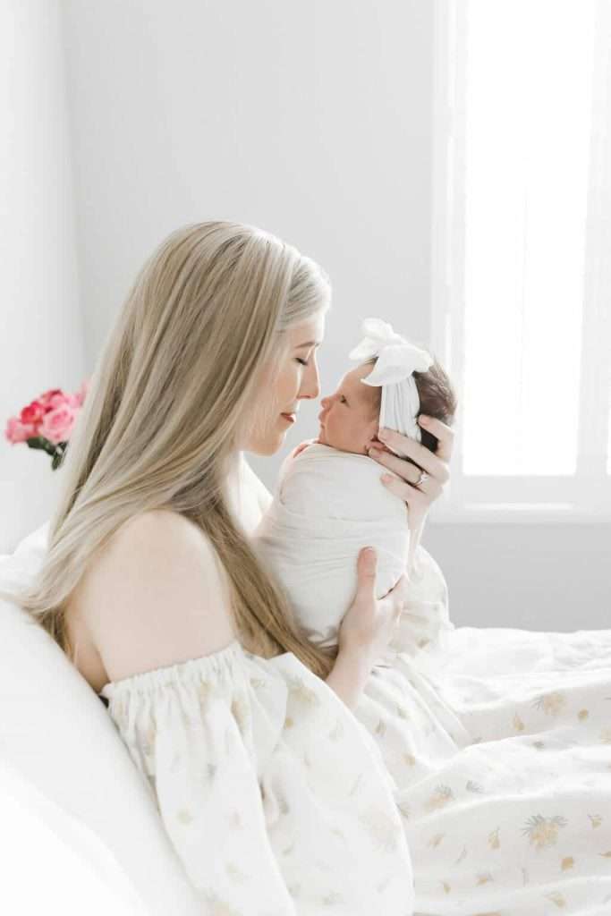 Woman with long blonde hair holds a swaddled newborn wearing a white headband, sitting on a bed with soft lighting and pink flowers in the background.