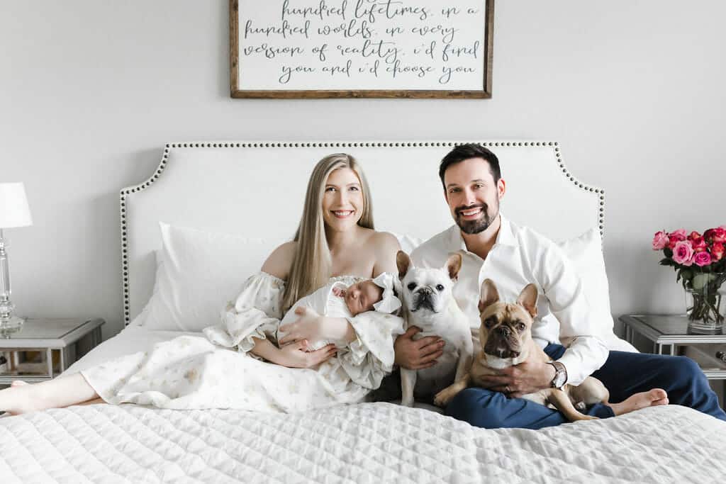 A woman, man, newborn baby, and two French Bulldogs sit together on a white bed. A framed quote hangs above the bed, and a vase of pink roses is on a bedside table.