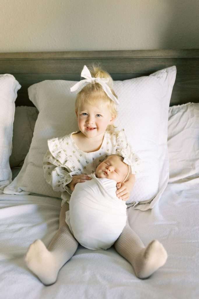 A young child with a white bow sits on a bed, smiling and holding a swaddled newborn baby in their lap.