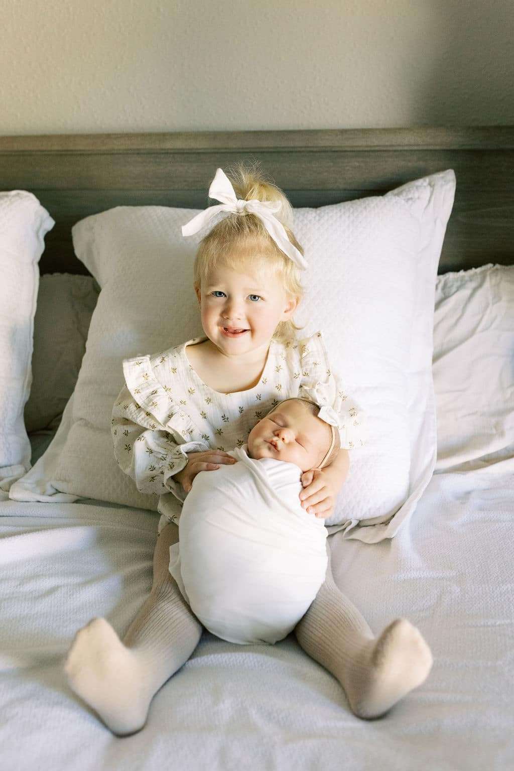 A young child with a white bow sits on a bed, smiling and holding a swaddled newborn baby in their lap.