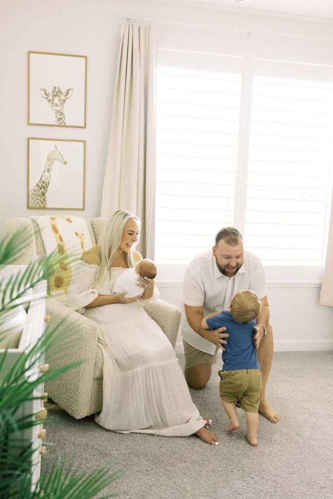 A woman holding a baby sits in an armchair while a man kneels, smiling at a young child who is walking toward him in a bright, cozy room with giraffe art on the wall.