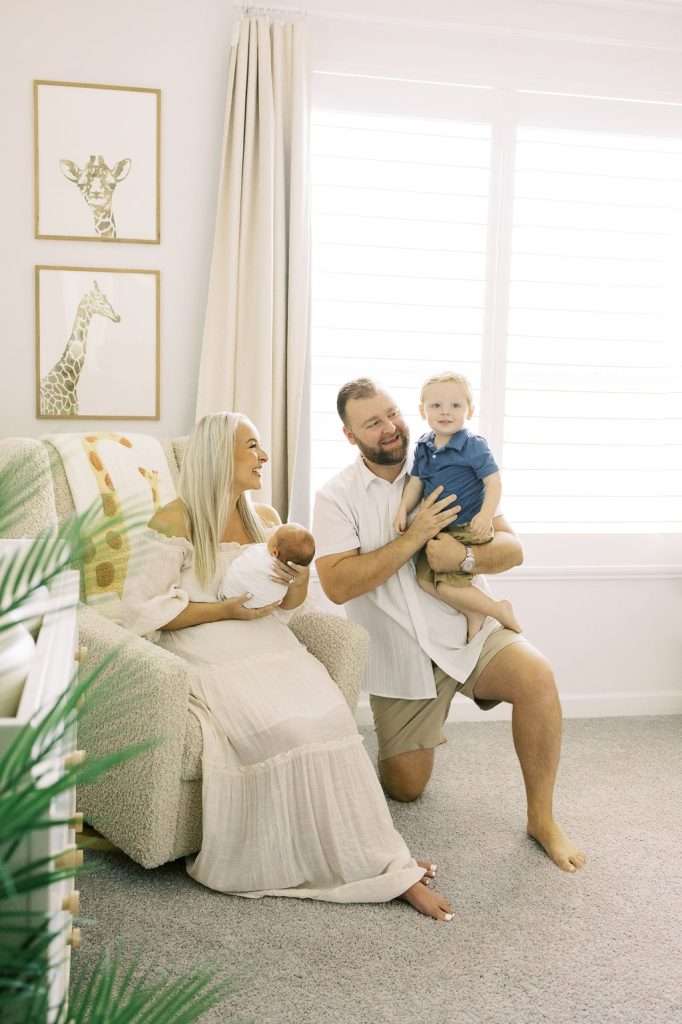 A woman sits holding a baby in a nursery chair while a man kneels beside her, holding a toddler. Two giraffe pictures hang on the wall behind them.