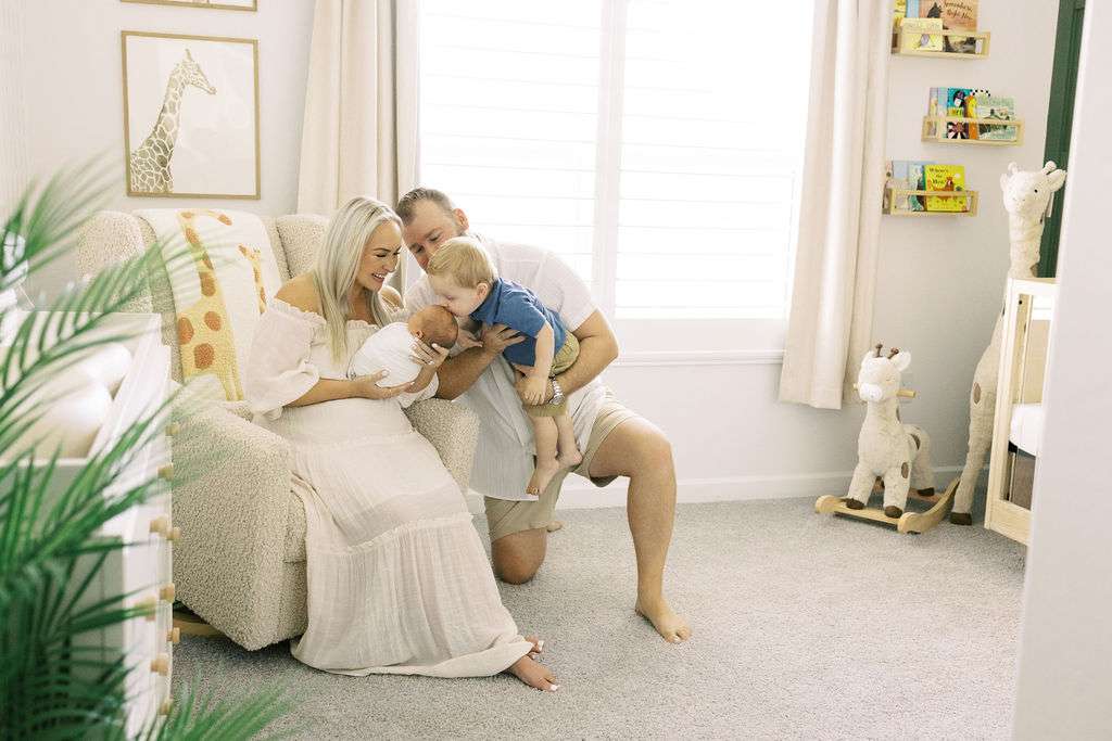 A woman sits in a nursery holding a baby, while a man kneels and helps a toddler kiss the baby. The room has soft lighting, animal decor, and children's books on shelves.