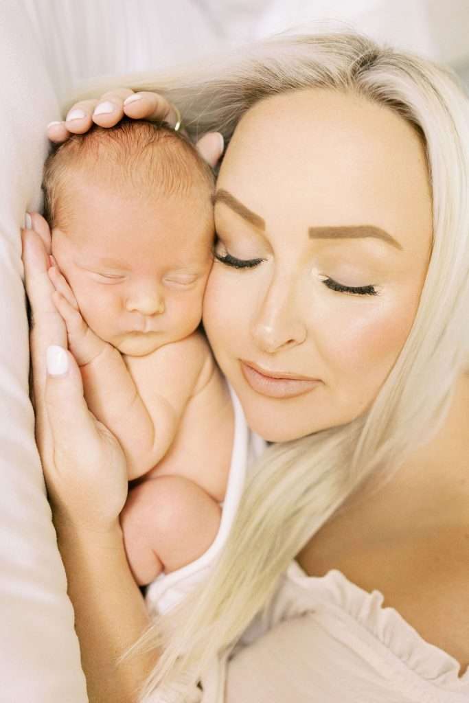 A woman with blonde hair lays closely beside a newborn baby, both with eyes closed, touching faces and hands in a calm, intimate moment.