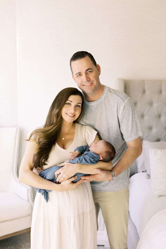 A woman and man stand close together in a bedroom, smiling at the camera while the woman cradles a sleeping newborn baby wrapped in a blue blanket.