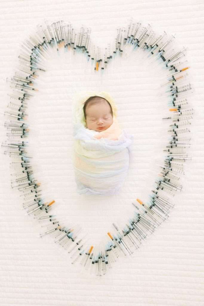 A newborn baby wrapped in a blanket lies in the center of a heart shape made from medical syringes on a white background.