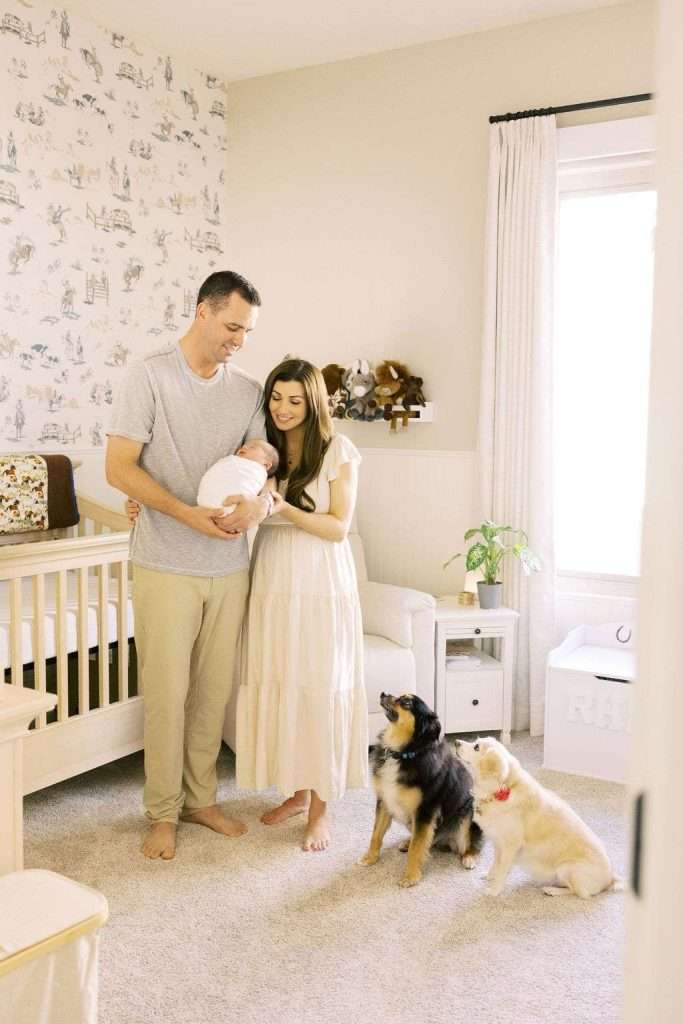 A man and woman stand in a nursery holding a baby, with two dogs sitting on the carpet in front of them.