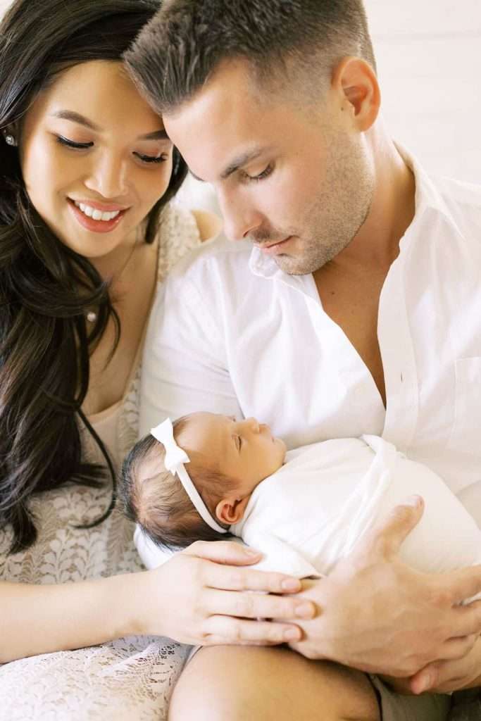A man and woman sit close together, smiling and looking lovingly at a newborn baby wrapped in a white blanket and wearing a white headband.