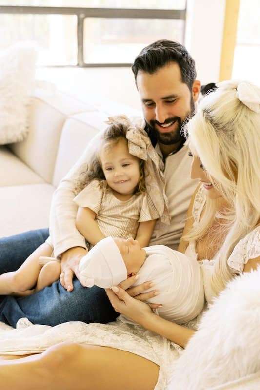 A family of four sits on a couch; the mother holds a newborn wrapped in a blanket, while the father and young daughter sit close, all smiling and looking at the baby.