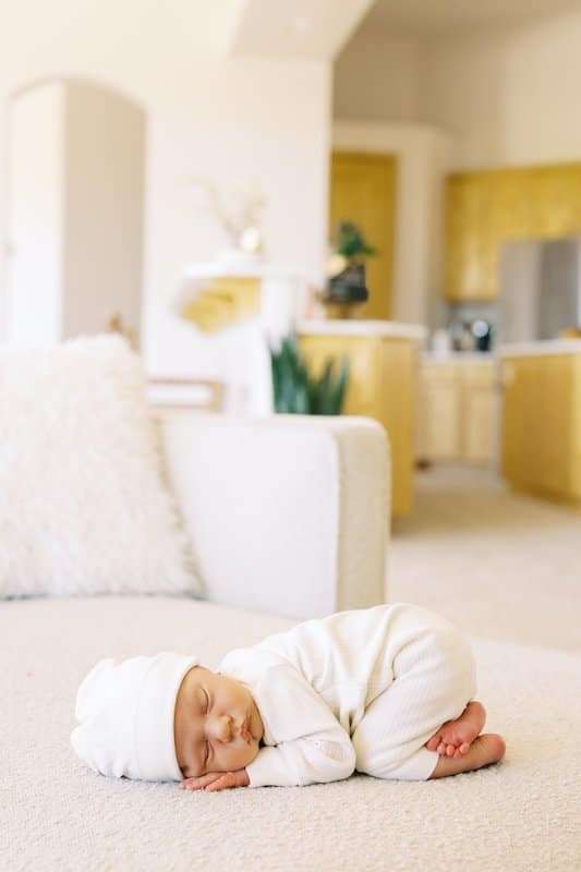 Newborn baby in white clothes and hat sleeping curled up on a light-colored couch in a bright, modern living room with a beige and yellow color scheme.