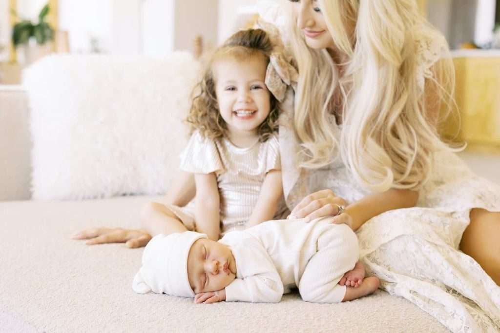 A woman sits on a couch with a smiling young girl and a sleeping baby dressed in white clothes and hat, lying on a soft surface.