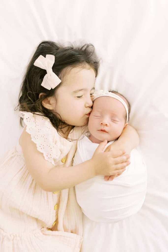 Young girl with a bow kisses and hugs a swaddled newborn baby on a white blanket.