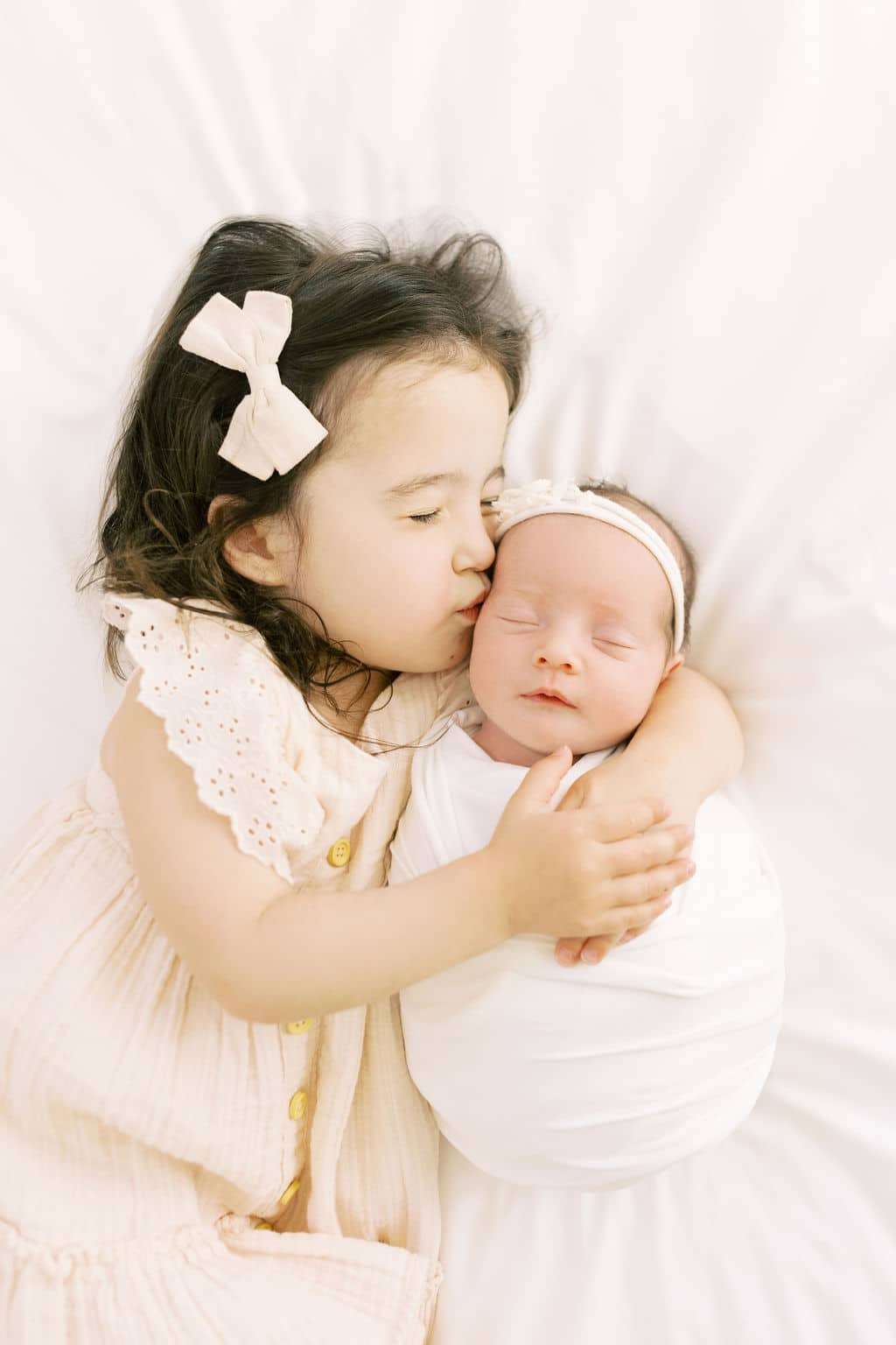 Young girl with a bow kisses and hugs a swaddled newborn baby on a white blanket.