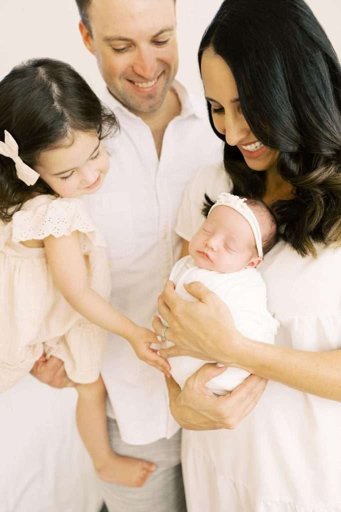 A woman holds a swaddled newborn while a man and a young girl stand close, all smiling and looking at the baby.