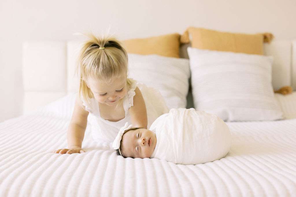 A young child in white looks at a swaddled baby lying on a white bed with pillows in the background.