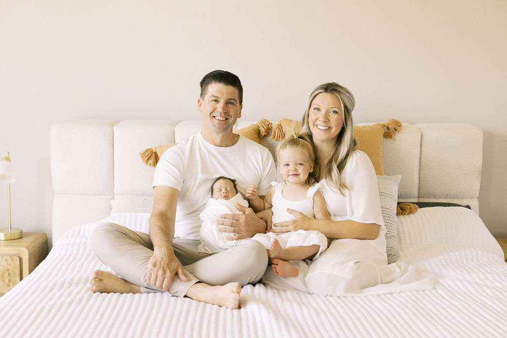 A family of four sits on a bed, with two adults and two young children, all dressed in light-colored clothing, smiling at the camera.