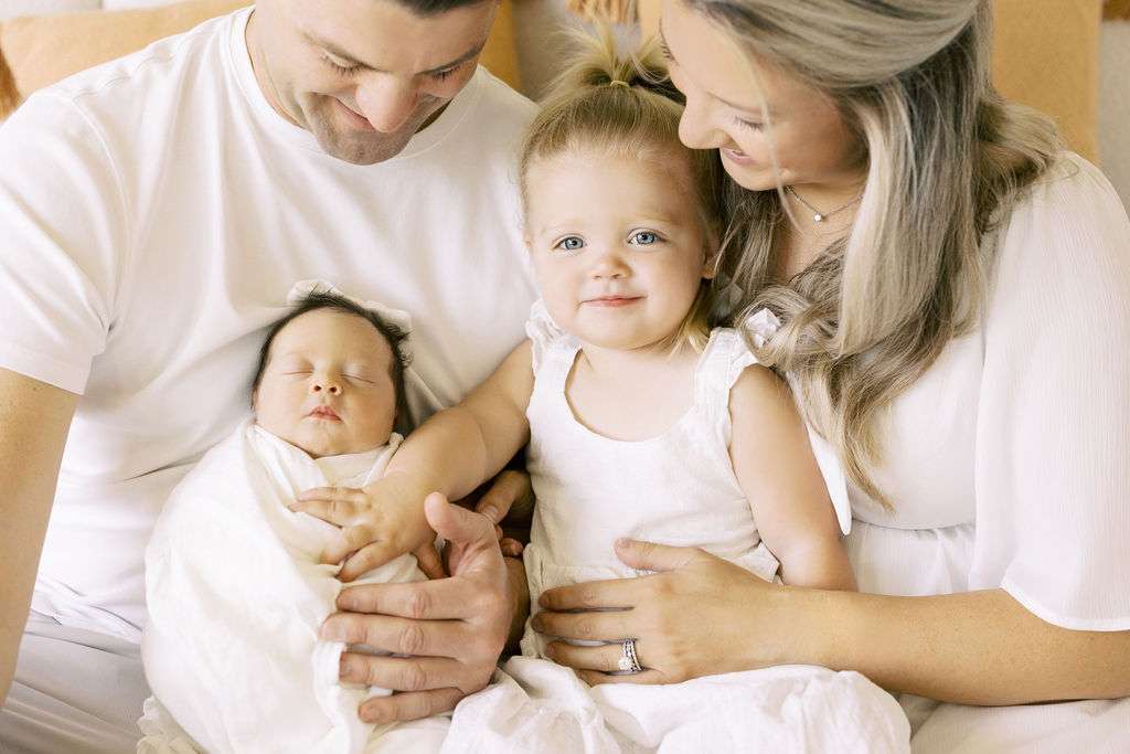 A man and woman sit close together, holding a sleeping baby and a smiling toddler dressed in white.