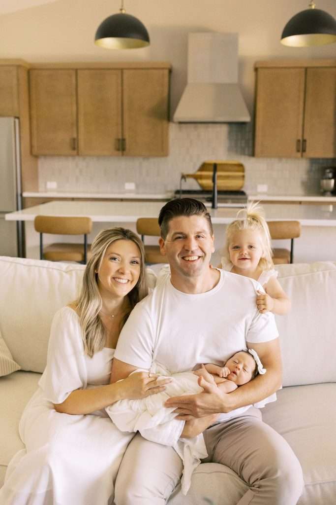 A family of four sits on a couch in a modern kitchen, with the parents holding a newborn and a toddler standing beside them.