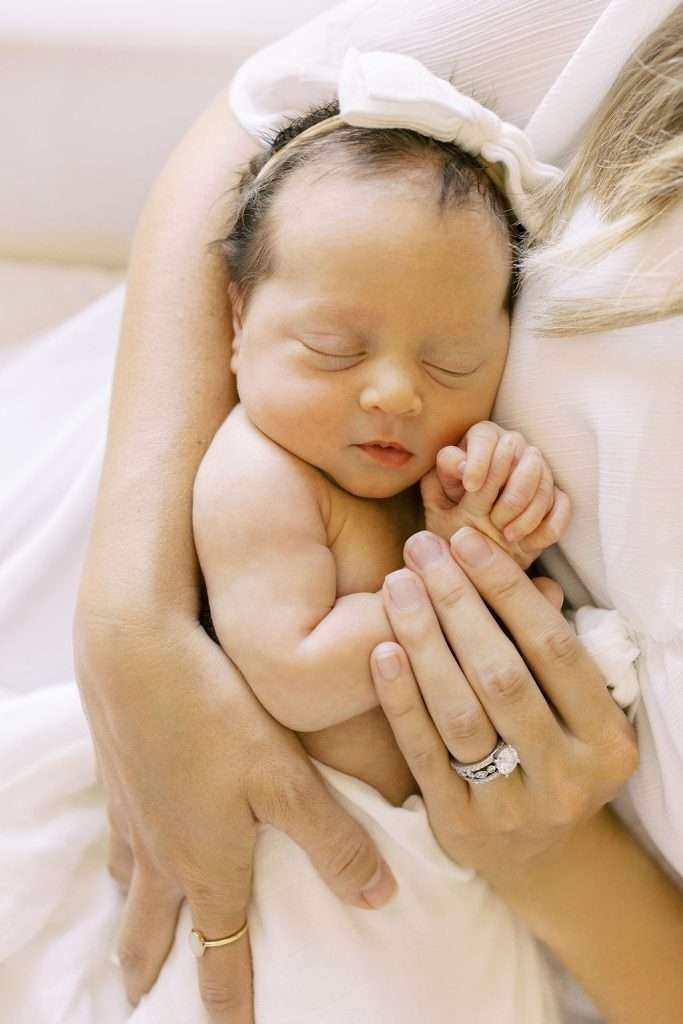 Adult cradling a sleeping newborn baby with a white bow headband. The adult’s hands are gently holding the baby, showing a wedding ring.