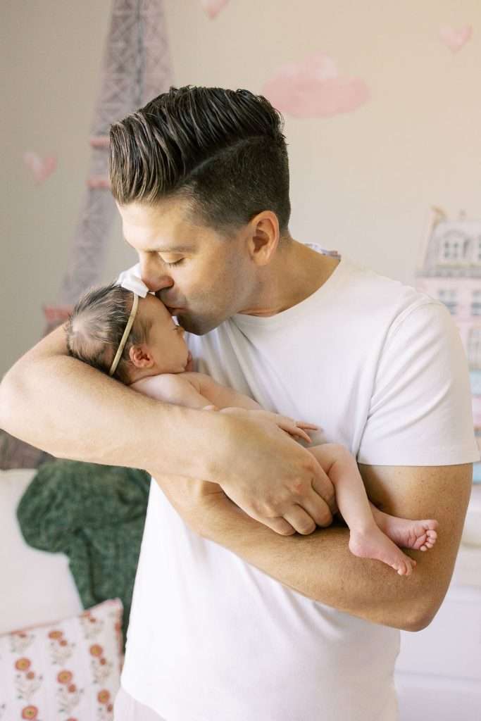 A man in a white shirt holds and kisses a newborn baby with a white bow headband in a softly decorated room.