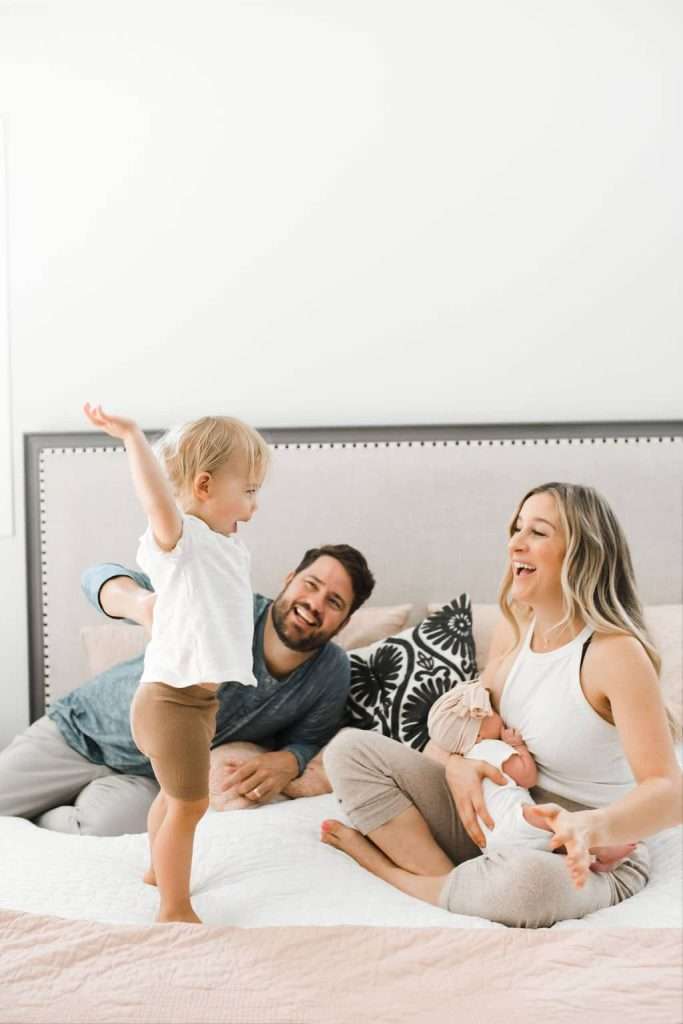 A toddler stands on a bed with arms outstretched as two adults smile nearby; one adult holds a baby while sitting on the bed.