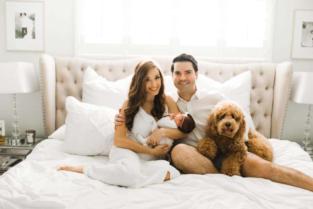 A couple sits on a bed holding a newborn baby, with a brown dog beside them. The setting is a bright, white bedroom with framed photos on the walls.