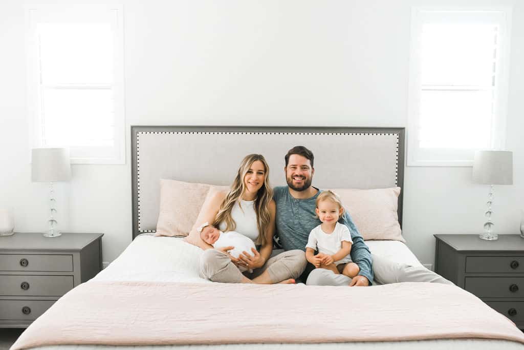 A family of four sits on a large bed in a bright bedroom; the mother holds a baby, the father and young child sit beside them, all smiling at the camera.