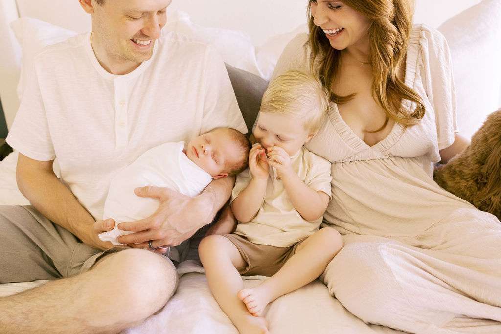 Two adults and two young children sit closely together on a bed. One adult holds a swaddled baby while the other child sits between the adults, smiling and touching their face.