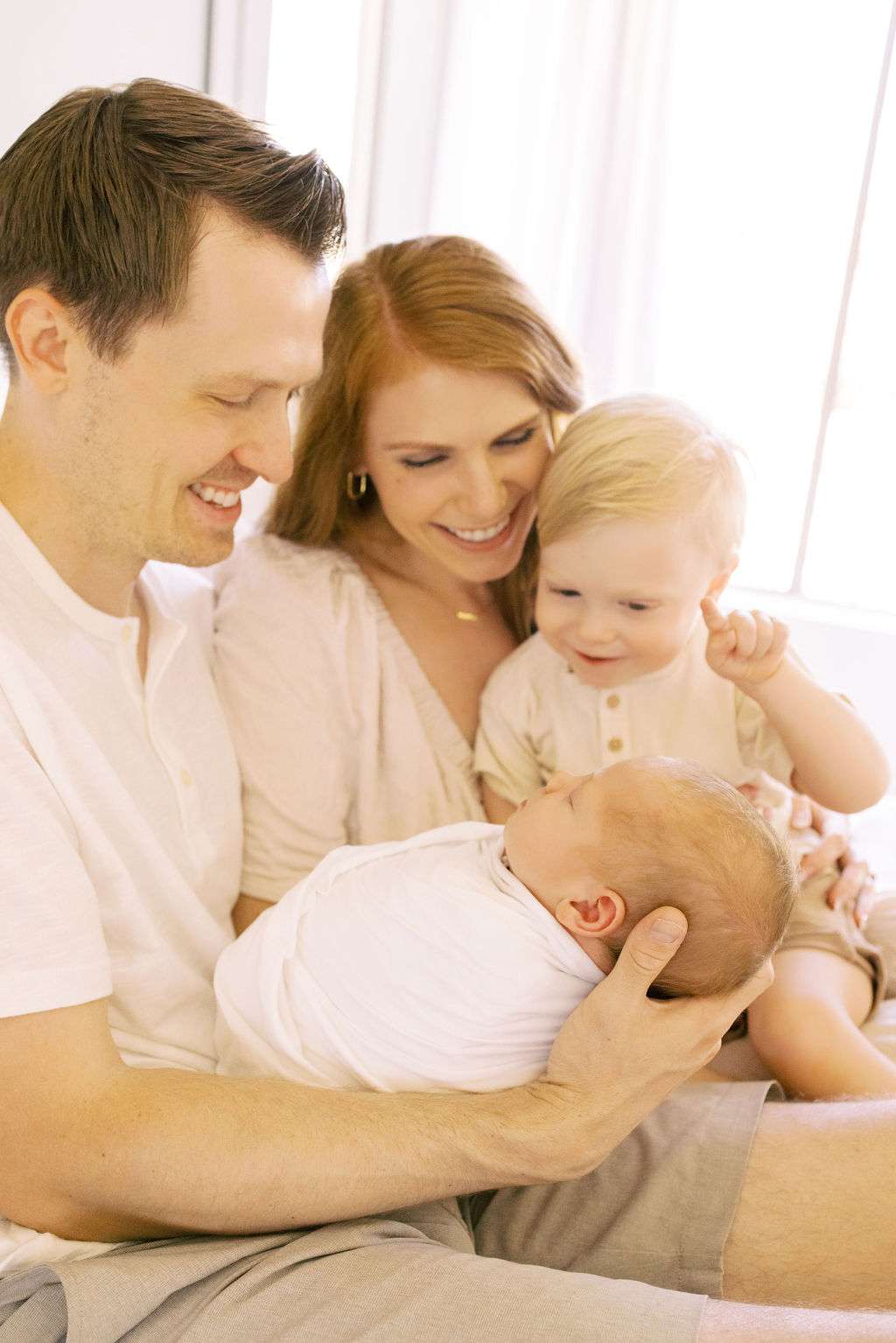 A man and woman smile while sitting with a toddler and holding a swaddled newborn baby in their arms by a window.