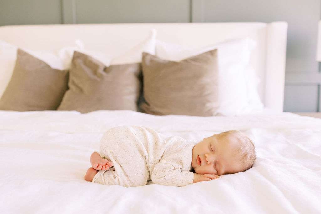 Newborn baby sleeping on a white bed, facing left, with head resting on hands and three pillows in the background.