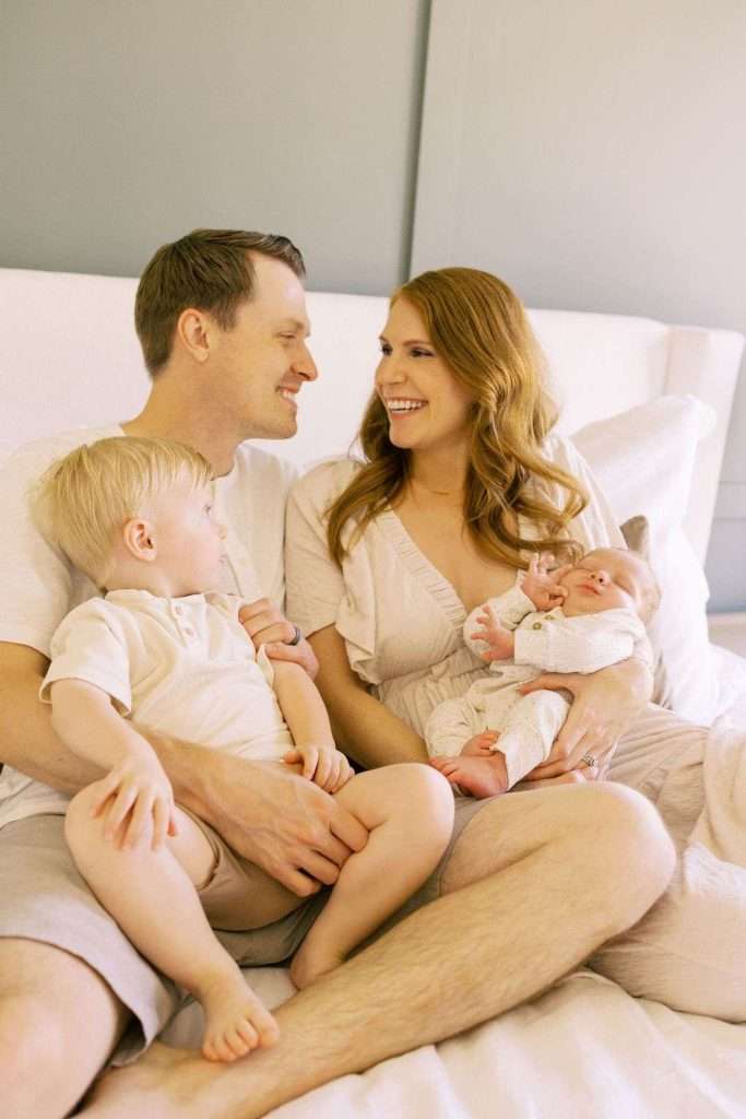 A family of four sits on a bed. Two adults smile at each other while a young child sits on one’s lap and a baby rests in the other's arms.