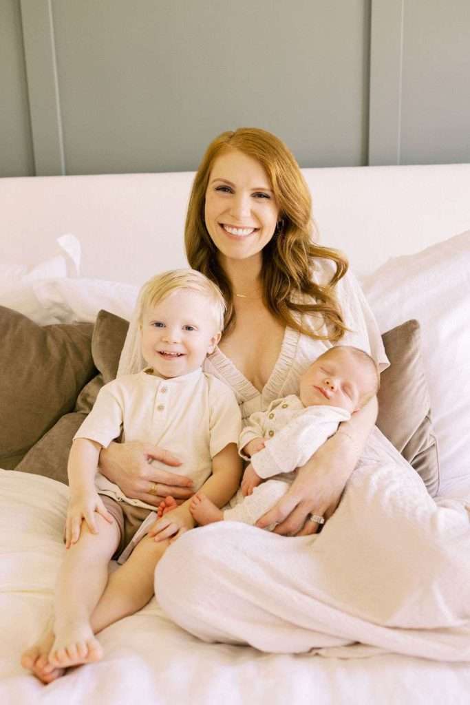 A woman sits on a bed holding a toddler and an infant, all dressed in light-colored clothing, smiling at the camera.
