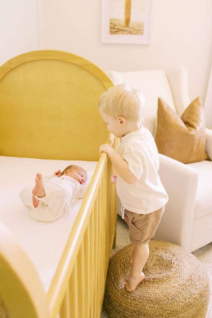 A toddler stands on a woven ottoman, looking into a crib at a baby lying on their back in a softly lit nursery.