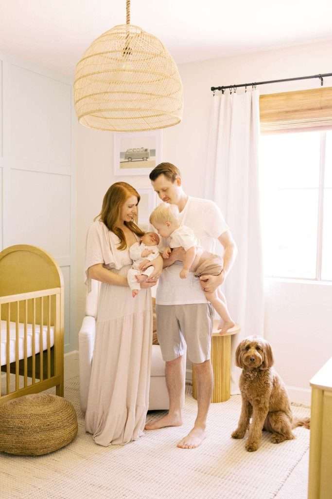 A man and woman stand in a nursery with two young children; a dog sits on the floor nearby. The room has soft lighting, a crib, chair, and woven decor.