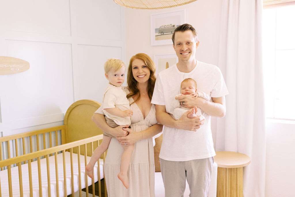 A man and woman stand in a nursery, smiling, with the man holding a baby and the woman holding a toddler. A crib and soft lighting are visible in the background.