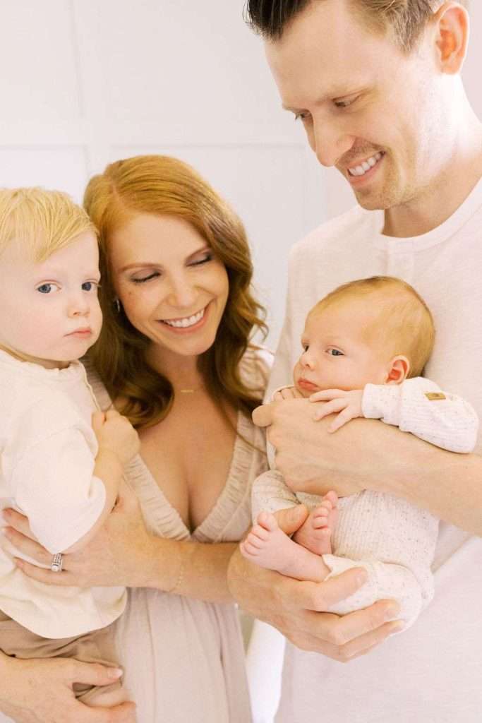A woman holding a toddler stands next to a man holding a newborn. They are all dressed in light-colored clothing and appear indoors.
