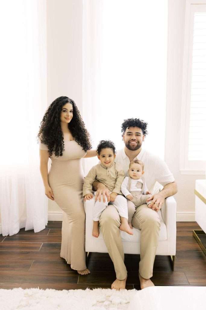 A woman stands next to a man seated in a chair, holding a baby and a young child. All four are dressed in neutral tones in a brightly lit room.