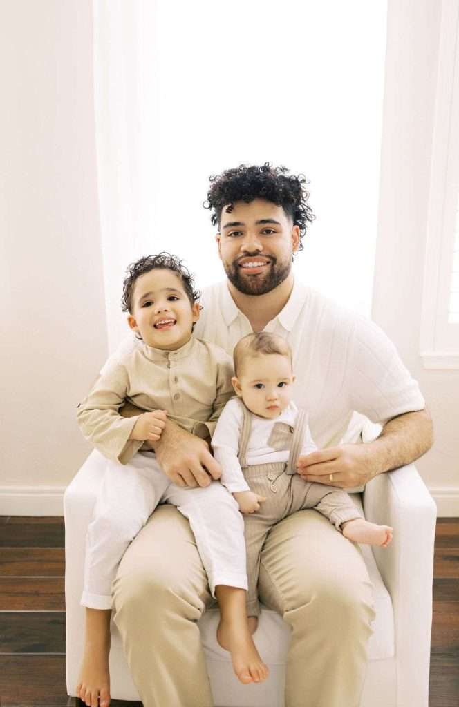 A man sits on a white chair holding two young children, both dressed in beige outfits, in a bright room with a large window behind them.
