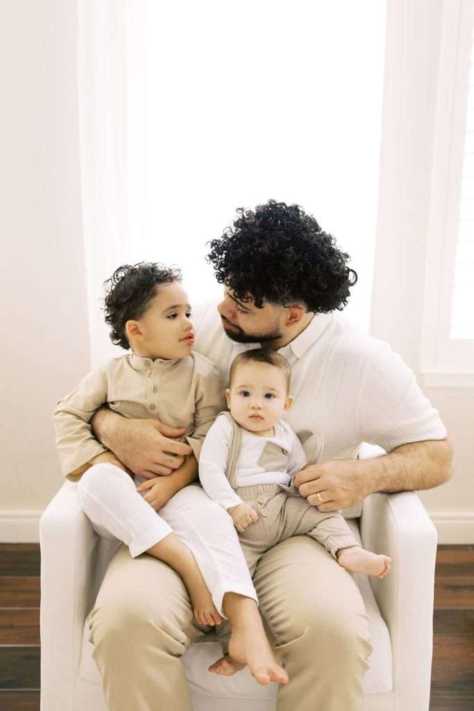 An adult sits in a white chair holding two young children on their lap, all wearing light-colored clothing, in a bright room with white walls and wood floors.