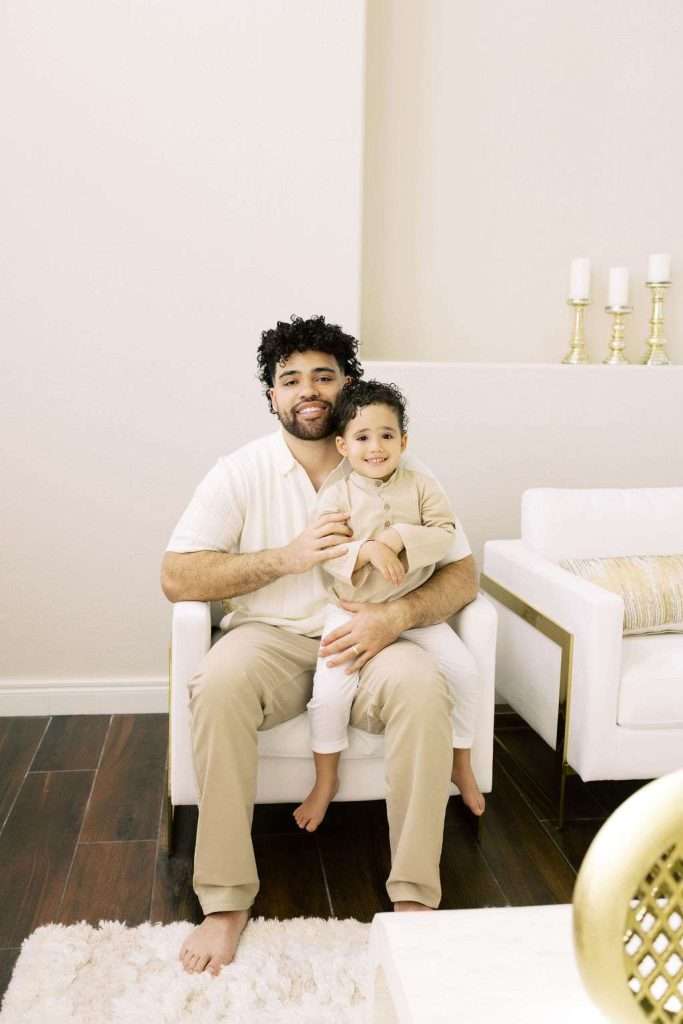 A man sits barefoot in a white armchair with a young child on his lap. Both are smiling and wear beige outfits. The room has white furniture, gold candle holders, and a light rug.