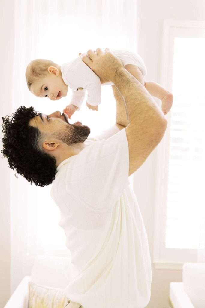 An adult lifts a baby overhead in a bright room, both dressed in white. The baby looks down, and the adult looks up, smiling.