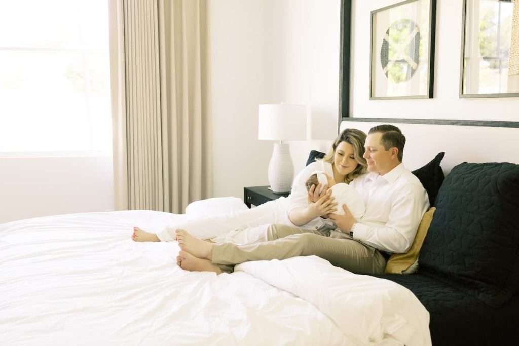 A couple sits on a bed holding a newborn baby, both looking at the child. The room is bright with white bedding, beige curtains, and framed art on the wall.