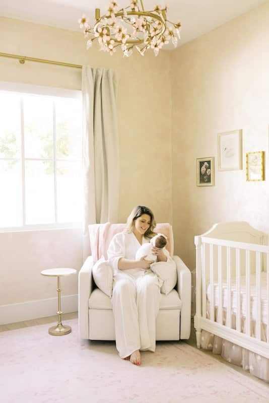 A woman in a white robe sits in a nursery, holding a baby. The room has a crib, soft lighting, framed pictures on the wall, and a window with curtains.